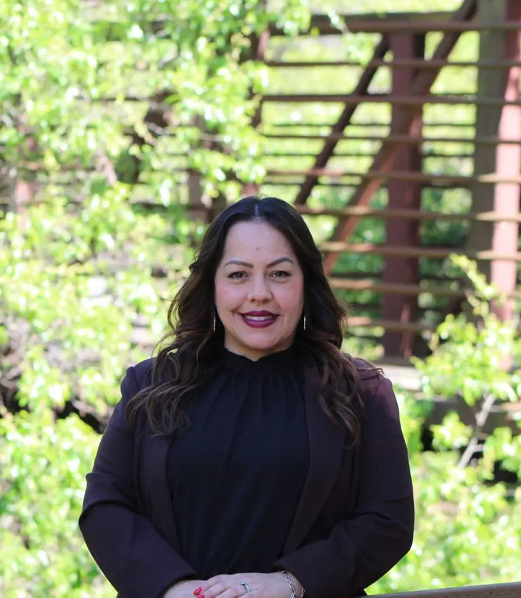 Nayeli in a black outfit standing outdoors with greenery and a bridge structure in the background